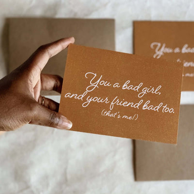hand holding orange card that has the words "You a bad girl, and your friend bad too. (that's me!)" in white cursive text.