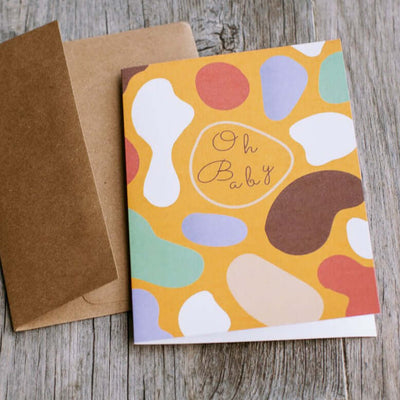 "Oh Baby Card" with colorful abstract illustration with that text "Oh Baby" in the middle written in a childish font showcased with the brown envelope it comes in