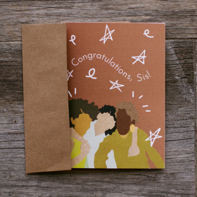 red card placed on a wooden backdrop that reads "congratulations sis" with an illustration of 3 people hugging and white stars.