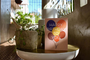 light pink Happy birthday card that reads "Happy birthday to you" in white text in front of a set of 6 different colored balloons. The card is on a side table with a small plant beside it.