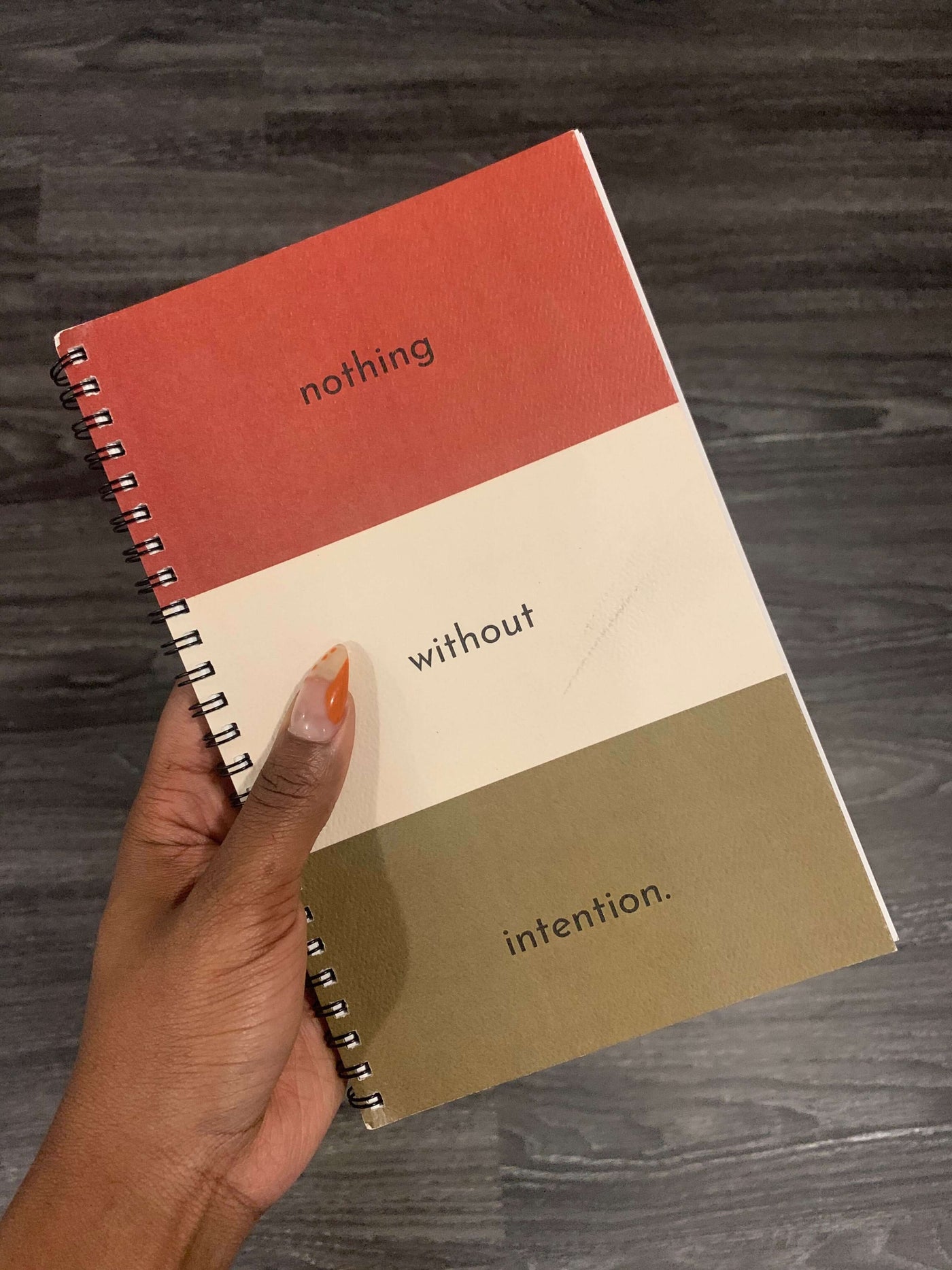 hand holding intention journal that is split into three equal colors of red white and greenish brown that read "nothing", "without", "intention" in each space on the cover