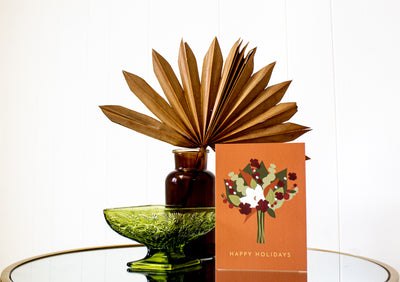 orange holiday floral greeting card that says happy holidays on it placed in front of a decorative plant.