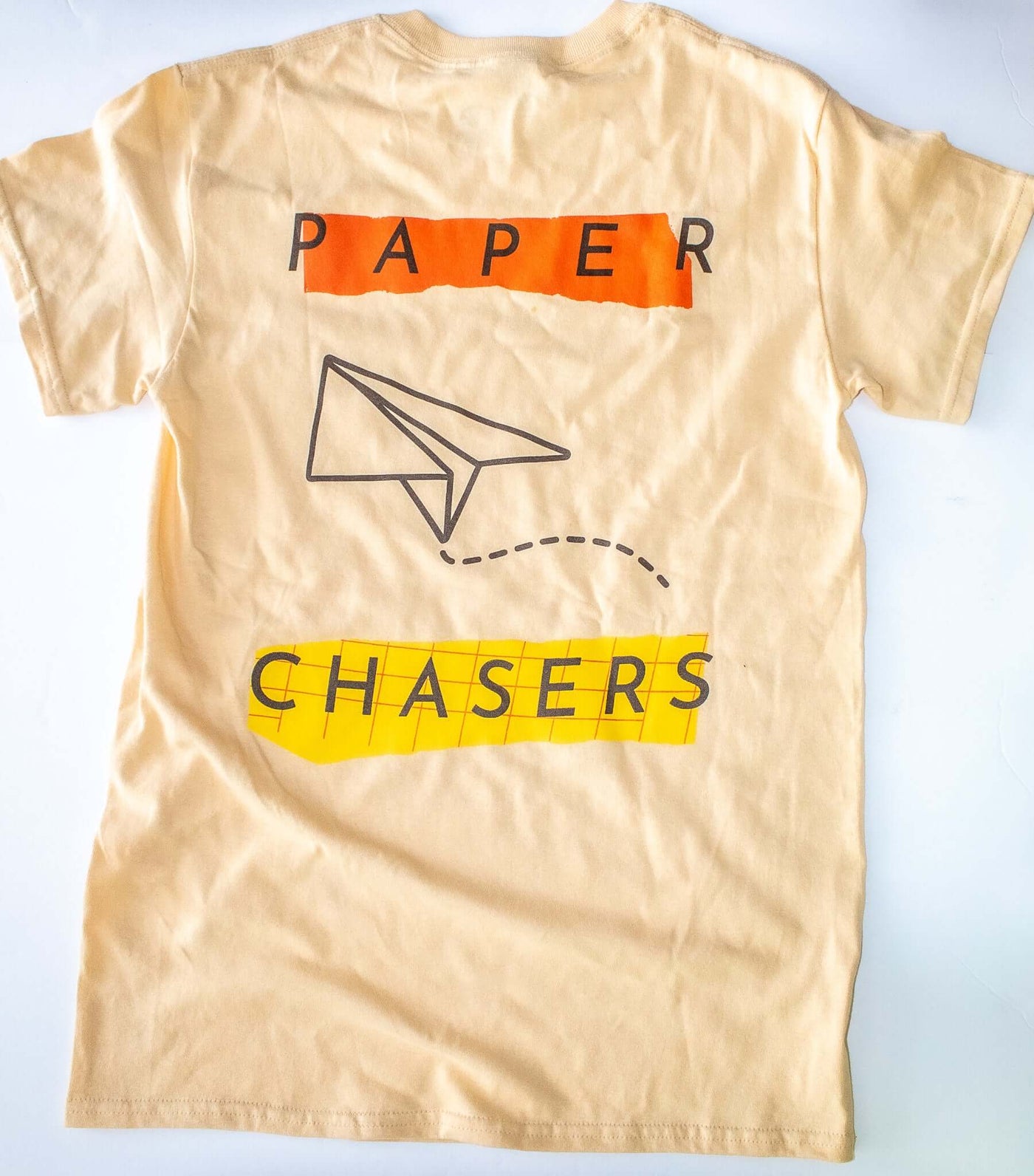 back of the tan colored "Paper Chasers T-Shirt" that has an illustration of a paper airplane in the middle.