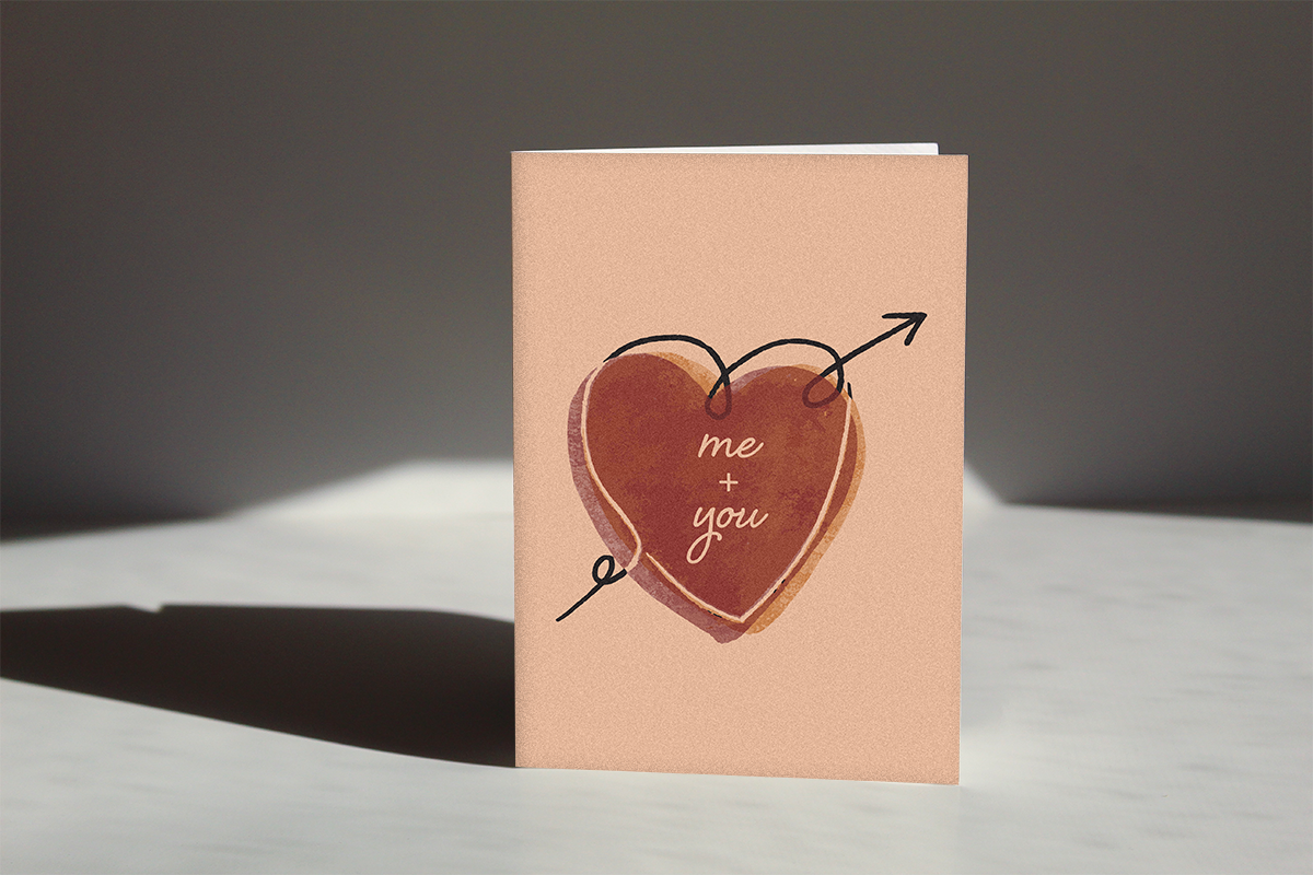 beige and pink colored "Me & You Romance Card" with a heart illustration and within the heart the text reads "Me + You" showcased on a counter in a spotlight