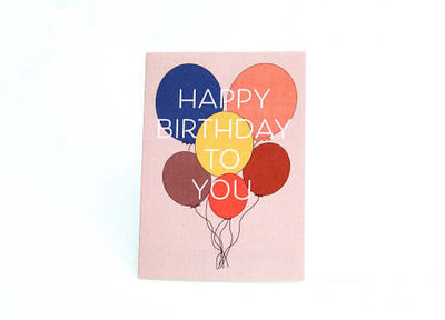 light pink Happy birthday card that reads "Happy birthday to you" in white text in front of a set of 6 different colored balloons.